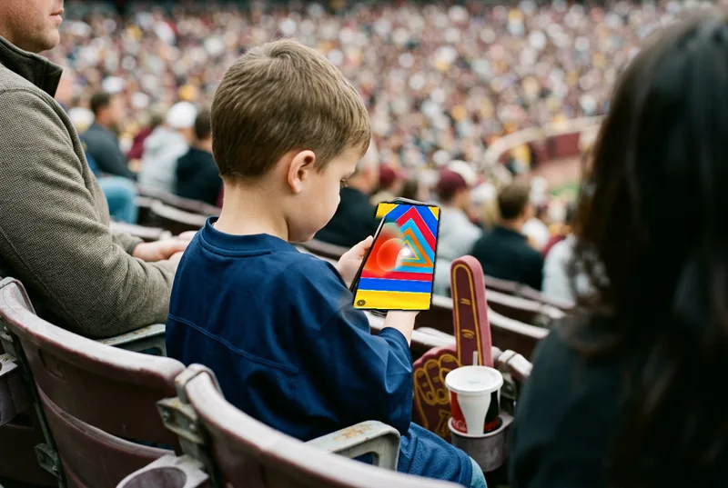 A boy in a plain blue jersey in stadium seats, holding a phone running Ondo while the game goes on behind him.